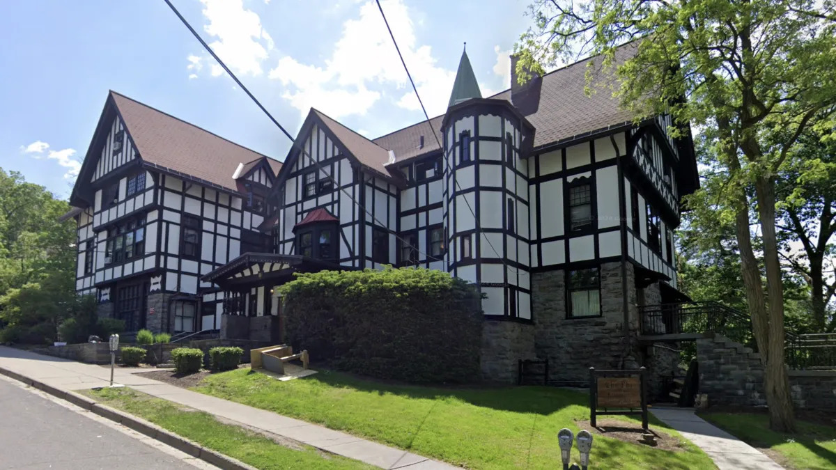 Exterior of a Tudor-style fraternity house on a tree-lined street.